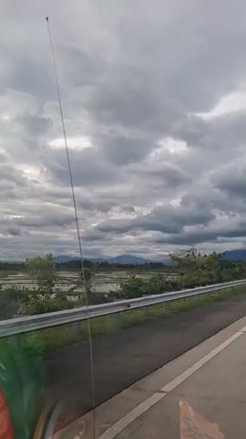 A vehicle is moving along a road with a silver guardrail next to it. Beyond the guardrail is a view of rice paddies, trees, and distant mountains. The sky is overcast with thick, grey clouds.  An antenna is visible, extending from the vehicle toward the sky. Part of the dashboard or interior of the vehicle is visible in the lower left of the frame.  A partially visible candy wrapper is in the lower right.
