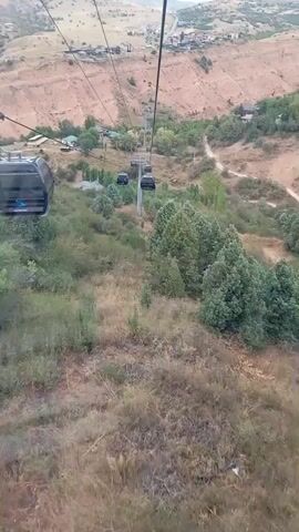 The video is a first-person perspective from a cable car ascending a mountain. The cable car is dark gray and has several windows. It is attached to a thick black cable that runs towards the top of the frame. Below, there is a lush green valley with scattered trees and bushes. A dirt road winds through the valley, and a few small buildings are visible. In the distance, the mountain slopes are steep and rocky, with some terraced areas suggesting human activity. The sky is overcast. The cable car appears to be moving at a moderate pace, and the camera angle is slightly tilted downwards.