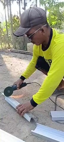 A man wearing a yellow long-sleeved shirt, black shorts, a dark baseball cap, and glasses is crouched on the ground outdoors. He is using a black and teal angle grinder to cut a piece of silver metal tubing, which is resting on the ground. Sparks are flying from the point of contact between the grinder and the metal. The ground is covered in dirt and loose gravel. In the background, there are trees and some construction materials, including more silver metal tubing. The man's hands are holding the angle grinder and supporting the metal tubing.