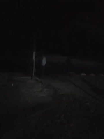 The video shows a dark outdoor scene at night, possibly a parking area. A person is initially standing near a pole, and as the camera pans, a motorcycle comes into view. A small, illuminated building is visible in the background, and there are other parked motorcycles nearby. The overall environment appears dimly lit.