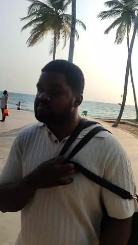 The video showcases a lively beach setting, probably in the tropics, with numerous palm trees lining the shore. The initial shot is of a man standing on a paved walkway, with the ocean and beachgoers visible in the background. As the video progresses, it pans to reveal a bustling scene of people walking, carrying items, and seemingly enjoying themselves. The presence of speakers suggests music is being played, contributing to the festive atmosphere. The focus shifts to two men dressed in matching colorful outfits, standing near a table. Overall, the video captures a vibrant and social beach environment.