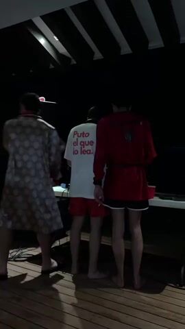 The video shows a group of young adults at a party, transitioning between indoor and outdoor settings. The first scene is on a dark wooden deck with three people standing near a table and a speaker. One person wears a patterned robe, while the others wear red shorts and robes, one red and one white. The white shirt has "Puto el que lo lea" printed on it. The next scene is indoors, showing people sitting on a couch. Two women are sitting together, one in a denim skirt, while a man in sunglasses lies next to them. There is a yellow plastic cup on a table. The last scene is a brief shot of a dark ceiling with lights as someone walks by. The music is energetic throughout.