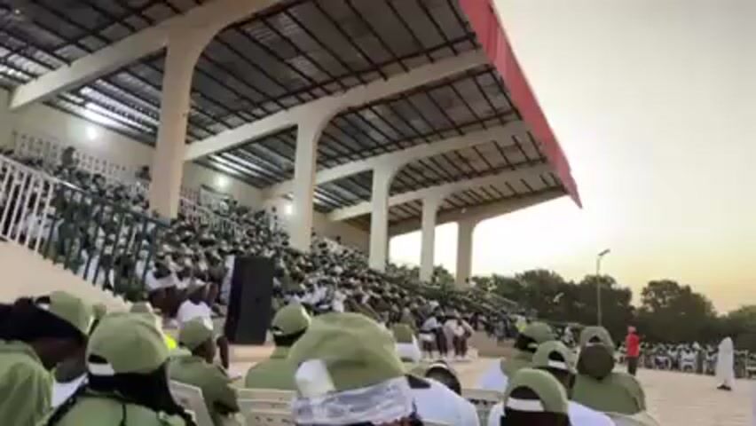 The video shows a large crowd of people, all wearing similar green and white outfits and green hats, seated in the stands of what appears to be a stadium. The stands are covered by a large metal roof, painted red along its edge and supported by white concrete pillars. The sky is a pale blue, suggesting it is either early morning or late afternoon. In the foreground, individuals in the same green attire are visible, some wearing plastic hair coverings. A speaker can be seen on the left side. The setting appears to be some sort of organized event or gathering.