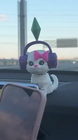 The video shows a bobblehead of a white cat with a pink bow on its head and purple headphones on. It also has a green Sims plumbob on top of its head. The bobblehead sits on the dashboard of a car, next to a cell phone in a pink case. The background shows a highway and a cloudy sky. The car is in motion, causing the bobblehead to wobble.