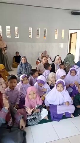The video shows a gathering of children and adults in a room with white walls and a tiled floor. The children, mostly young, are dressed in purple and pink hijabs and clothing. They are sitting on the floor in rows. Adults are seated behind them, also on the floor. Some of the adults are wearing hijabs. The room appears to be a community hall or a religious space. In the later part of the video, the scene changes to show decorated structures, possibly floats or displays, adorned with various items, including packaged goods and fabric. The overall atmosphere seems to be festive or celebratory.