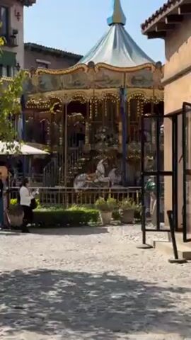 The video shows a carousel in an outdoor, cobblestone courtyard. Several people are walking around the carousel, some of whom appear to be a family. There are planters, outdoor seating with umbrellas, and a building beside the courtyard, with doors and windows visible. The carousel is spinning, with figures riding on it. The weather appears sunny and bright.