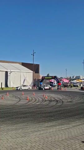 A drift racing event is underway on a sunny day. The video pans from a white tent and a drift car performing donuts on a paved track marked with orange cones to a large crowd of spectators gathered in tiered seating areas. The spectators, dressed in casual summer clothing, are watching the event with interest. Some are standing and cheering, while others are seated. A large, modern-looking structure with a metal roof provides shade for a portion of the seating. The overall atmosphere appears festive and energetic, suggesting a public event or competition.