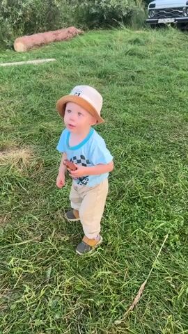 A young child wearing a light blue t-shirt, tan pants, a white bucket hat with a number on it, and dark shoes stands in a grassy field. The child holds a brown object in their right hand. The child turns their head to the left, then to the right, and then to the left again. In the background, a large log lies on the grass to the left, and the rear of a white vehicle is visible on the right.