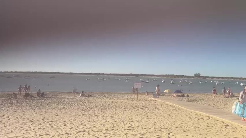 The video shows a wide shot of a sandy beach on a clear, sunny day. The ocean is calm with numerous boats anchored offshore. A wooden walkway extends from the foreground into the scene. Several people are visible on the beach, some walking, others sitting or lying down. A few are gathered near the water's edge. A sign is posted on the beach.
