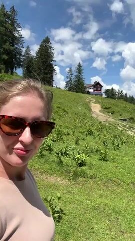 A person wearing sunglasses is visible on the left side of the frame. The video pans across a green, grassy mountain slope with scattered trees. In the background, there are mountains, a blue sky with white clouds, and a building on a hilltop. Further into the scene, a person is pushing a stroller on a gravel path. Several cows are grazing on the hillside near a fence and a small wooden structure. The camera continues to pan, showing more of the mountain landscape and the path.