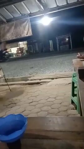 The video opens with a view of a dimly lit outdoor area at night, possibly a market stall or food stand. The ground is covered in gravel and hexagonal paving stones. A blue plastic bowl sits in the foreground. In the background, there are structures with lights and awnings, and people can be seen preparing food. One person, wearing a light-colored shirt and dark pants, is bent over, tending to something on a table. Another person, wearing a pink headscarf and a white shirt, stands near a counter. Several bowls of various colors, including red and green, are visible on tables. A glass of amber-colored liquid with a straw is also on a table in the foreground, and a hand is resting near it. The overall scene suggests a casual, outdoor dining or food service environment.