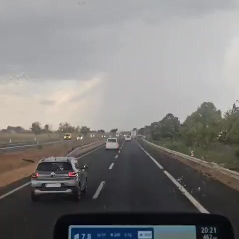 The video shows a view from inside a vehicle on a highway. The sky is overcast, and there are streaks of rain on the windshield. Several cars and trucks are visible on the road ahead, traveling in the same direction. The road is asphalt, with white lane markings. Trees and fields are visible on either side of the highway. The timecode indicates the video is about 5 seconds long.