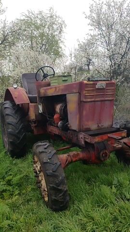 The video shows a rusty, maroon-colored tractor in a grassy field. The tractor has large, worn tires, an exposed engine, and a partially disassembled frame. There's a green crate on the hood and a single headlight. Behind the tractor are trees with white blossoms, suggesting it's springtime. The engine appears to be running.