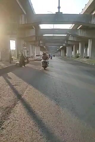 A video shows a person riding a scooter under a highway overpass. The road is asphalt and appears to be in a developing or industrial area. There are several other vehicles and people visible on the side of the road. The sun is shining, casting shadows.
