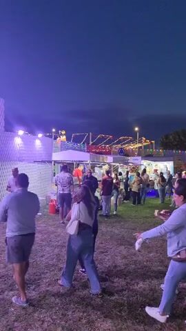 The video shows an outdoor event at dusk. In the foreground, several people are walking and standing on a grassy field. Some of them are holding drinks. In the background, there are various structures including tents, a Ferris wheel, and food stalls, all illuminated with colorful lights. The sky is a deep blue, indicating the transition from day to night.