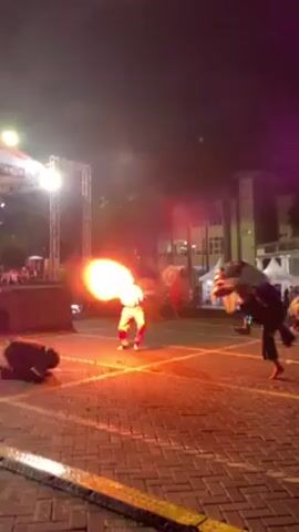 Here's a concise description of the video:

The video shows an outdoor performance at night. A person in a red jumpsuit is performing a fire-breathing act, creating a large burst of flames. Other performers are present, including one person in a monkey costume crawling on the ground and another wearing a large, elaborate headpiece dancing. A stage is set up in the background with lighting and a banner, and several white tents are visible around the perimeter. The performance takes place on a brick-paved area marked with yellow lines.
