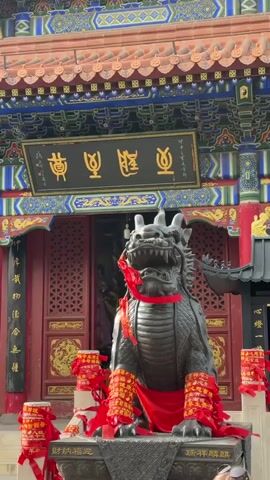 The video shows a statue of a dragon-like creature outside of a Chinese building. The building has a red and gold ornate door, with a sign above it in Chinese characters. The dragon statue is gray and has a red cloth draped over it. There are also several red and gold cylindrical objects decorated with ribbons around the base of the statue. The building’s eaves are intricately decorated with blue, gold, and red designs. Some tourists can be seen at the very bottom of the frame.