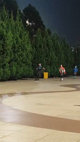 A group of three individuals are rollerblading on a paved outdoor area at night. Two of them are in the foreground, one in a black and grey shirt and dark pants, and another in a red and white jacket. The third individual, wearing a blue jacket, is further in the background on the right. A dense line of green trees forms a backdrop behind them. The ground is a light-colored paving stone with a curved, darker section in the middle. The lighting suggests artificial illumination and the presence of a moon partially obscured by trees.