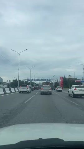 The video shows a view from inside a vehicle, looking forward. The vehicle is on a multi-lane road, with other cars visible in front and to the sides. There is an overpass ahead with large blue letters spelling "ВЛАДИКАВКАЗ", which appears to be a city or region name. To the right of the road, there is a gas station with a red and white sign displaying prices. The sky is cloudy, and the overall atmosphere appears to be overcast. The traffic appears to be moving slowly.