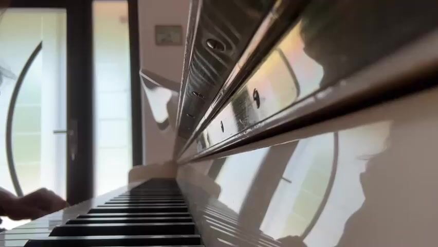 This media file captures a close-up, low-angle view of someone playing a white upright piano. The focus is on the piano keys and the player's hands, blurred by motion, as they strike the keys. The polished surface of the piano reflects the surrounding environment, including what appear to be large windows with a grid pattern, suggesting an interior setting. Sunlight streams in from the left, casting highlights on the piano and creating a warm, inviting atmosphere. The background is somewhat out of focus but shows a doorway with a handle and a small framed picture on the wall, providing hints of a domestic or possibly an institutional space like a music room or a living area. The overall mood is peaceful and contemplative, evoked by the act of playing the piano and the soft lighting. The time of day is suggested to be daytime due to the natural light. There is no visible text in the media file.