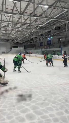 The video shows an ice hockey game in an indoor arena. The camera is positioned behind the goalie net, looking out onto the ice. Players in green and orange jerseys are skating and playing. The green-jerseyed goalie is in his net, with a player in a green jersey skating towards the net. Another player in an orange jersey is also near the net. The ice surface appears wet in places, with visible puck marks. Spectators are visible in the background in the stands. The arena has a metal roof structure with lights and banners. The scoreboard is visible in the background. The action appears to be in the defensive zone.