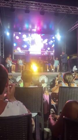 The video shows an outdoor stage at night, illuminated by bright lights and a large screen displaying colorful graphics. On stage, several people are seated in chairs, and one person is standing and holding a microphone, appearing to be a host or performer. The seated individuals are facing the audience. In front of the stage, an audience is seated in chairs, with their backs to the camera, watching the performance. The host is speaking into the microphone, and the audience is looking towards the stage.