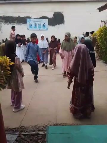 A group of children, mostly girls wearing hijabs, play a game outside on a paved area. They are gathered in a circle or loose formation, moving around and interacting with each other.  A boy in a blue shirt appears to be stepping over a rope or similar object.  In the background, other people are seated on the ground. A white building with a red tiled roof forms the backdrop of the scene.  A banner with images and text hangs on the wall of the building. There are plants and flowers around the perimeter of the playing area. The overall atmosphere is lively and playful.