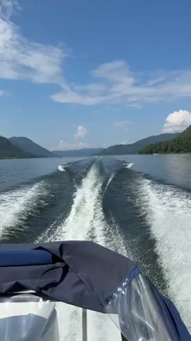 The video shows a view from the back of a boat moving on a large lake. The camera perspective is looking back towards the wake of the boat. The water is dark blue and shows a significant white wake spreading out behind the vessel. In the distance, there are other boats also moving on the water, creating their own wakes. The lake is surrounded by lush green, tree-covered mountains. The sky is a clear blue with some fluffy white clouds. The overall scene depicts a bright, sunny day on a body of water with mountainous scenery.
