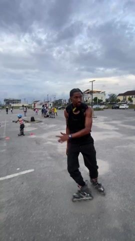 The video shows a man roller skating in an outdoor parking lot with a group of children practicing around cones in the background. The man, who is Black, wears a black tank top, black pants, a black vest with multiple pockets, and inline skates. A green helmet hangs from his backpack. He skates in a slow, controlled manner, looking around the parking lot. The sky is cloudy and there are buildings visible in the distance.