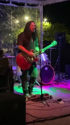 A rock band plays live music on a stage outdoors at night. The lead singer and guitarist stands front and center, playing a red electric guitar and singing into a microphone. The drummer is behind him, and two other guitarists flank him on either side. Stage lighting casts green, pink, and blue hues. Cables and equipment are strewn across the stage. The outdoor venue is lit by string lights and streetlights.