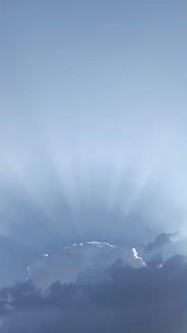 The video shows a bright sky with a cloud formation. Rays of light emanate from behind the clouds, creating a crepuscular effect. The clouds are illuminated from behind, with some appearing white and bright, while others are dark and shadowed. The sky is clear and blue, with no other objects or people visible.