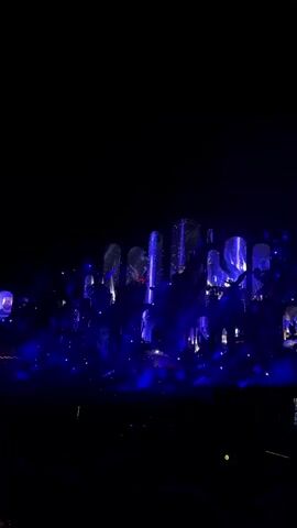The video shows a dark scene illuminated by bright blue lights. There appear to be many people gathered, suggested by the crowd-like density and scattered light sources. In the background, large, abstract structures or sculptures are visible, also lit with blue and white lights. Some of these structures have reflective surfaces, creating a shimmering effect. The overall atmosphere suggests a concert, festival, or other large-scale event.