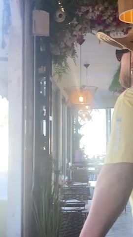 The video begins with a view of a cafe's exterior, showing an outdoor seating area with tables and chairs. A bicycle is parked near a wall. The camera then pans to the right, revealing the interior of the cafe or a covered outdoor seating area. A man is seated at a table with a cup of coffee and a slice of lemon. He is wearing a white t-shirt and has his hand covering his face. In the background, there are street signs and trees. The video briefly shows a portion of another person's arm and hand before transitioning back to the exterior view. The overall scene appears to be a relaxed, daytime setting at a cafe.