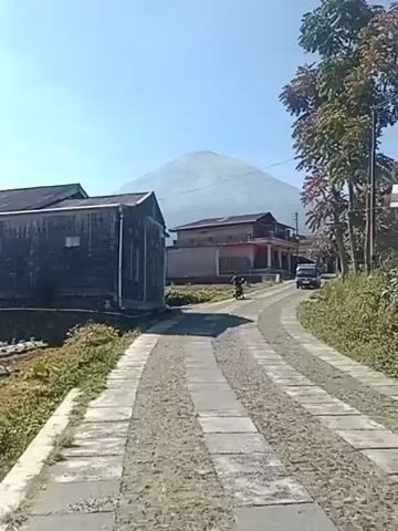 The video shows a narrow, unpaved road with concrete slabs running down the middle. On the left is a dark, weathered building with a single window and what appears to be a gutter at its base. To the right of the road, a small agricultural field is visible, bordered by a low brick wall. Further down the road, there are several houses, one of which has a red-tiled roof and balconies. A motorcycle with a rider is moving away from the camera, and a dark-colored vehicle is parked in front of one of the houses. In the background, a large, light-colored mountain dominates the horizon under a clear blue sky. The overall impression is of a rural village setting on a sunny day.