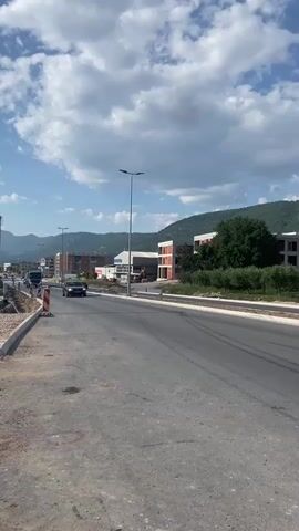 A truck is driving on a road with a car following behind it. There are buildings and trees on the side of the road, and mountains in the background. The sky is blue with some clouds.