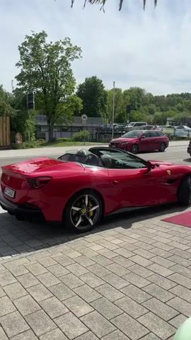 The video shows a red Ferrari Portofino parked on a gray stone-paved surface. The top is down, and two people are sitting inside. The car is parked near a building with a red carpet leading to the entrance. A red Peugeot is parked on the street behind the Ferrari. There are other cars parked further down the street. A large green tree is visible on the left side of the frame. The sky is mostly clear with some clouds.