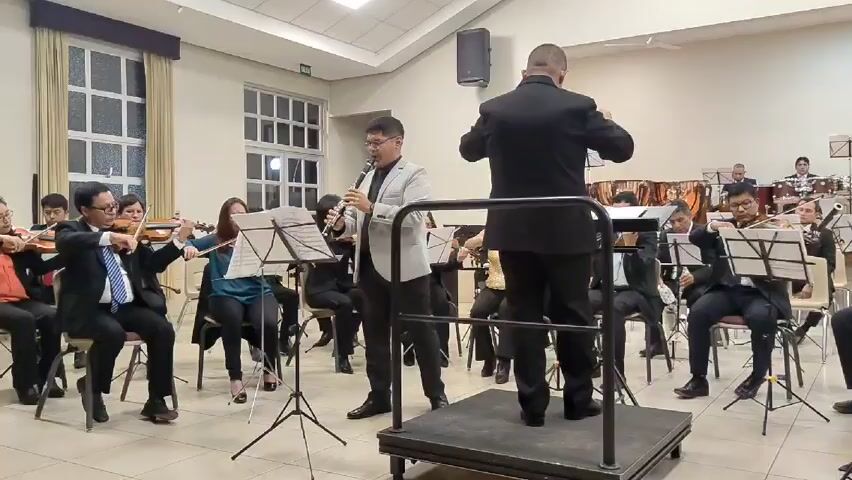 An orchestra is performing in a room. There are at least 10 musicians playing violins while seated, all of them wearing dark suits. A man in a grey blazer is standing in front, playing a clarinet. Another man in a black suit is standing on a small platform with a railing, facing away from the camera and conducting the orchestra. There are music stands in front of each musician. In the background, there are windows, a speaker on a stand, and additional musicians playing percussion instruments. The room is well-lit and appears to be a hall or community center.