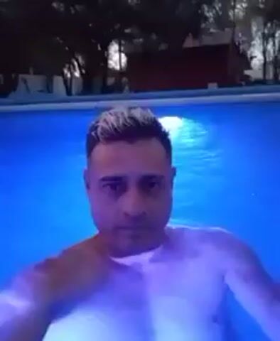 A medium shot shows a man in a pool, illuminated by a light under the water, looking at the camera. The water changes from blue to purple to green. The man has short, light-colored hair with darker roots. He appears shirtless. In the background, there are trees and a small building.
