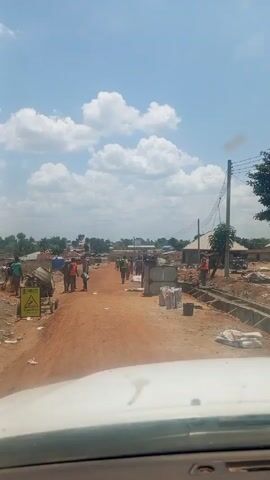 From the perspective of a vehicle driving down a dirt road, a construction crew is visible working on the road. There are about 10-12 people, dressed in work clothes and some wearing safety vests. They are grouped along the road, some standing, some carrying items, and some near equipment like a cement mixer. Large rectangular blocks and bags of materials are visible on the side of the road, next to a caution sign. The road runs through a rural area with simple houses and some trees. The sky is blue with scattered clouds.