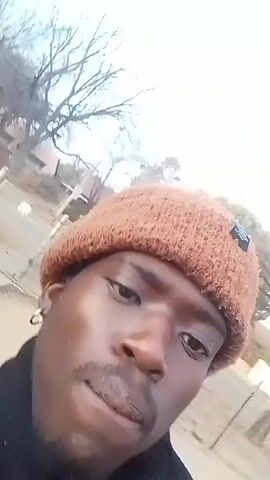 A young Black man wearing a brown knitted beanie, a black jacket, and a Christmas-themed sweater, films himself outdoors in an overcast environment. He shows his teeth and briefly yawns. In the background, there is what appears to be a residential area with bare trees and fences. Music can be heard.
