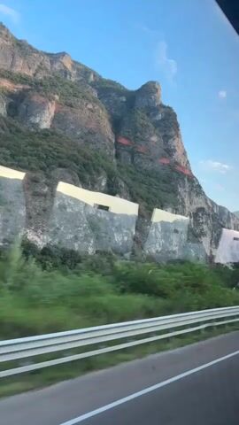 A video shows a mountain face with reddish-brown strata visible against gray rock. Several large, white, angular structures are built into the mountainside, some with dark rectangular openings. Green vegetation grows at the base of the mountain and also on the slopes between the structures. In the foreground, a highway guardrail is visible, indicating the perspective is from a moving vehicle. The sky is clear and blue.