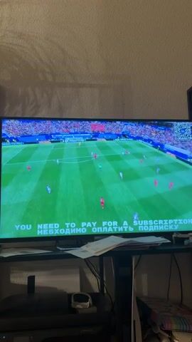 A television screen displays a soccer match. The game is being played on a green field with white lines. Players in red and white uniforms are on the field. A message at the bottom of the screen reads, "YOU NEED TO PAY FOR A SUBSCRIPTION." The background appears to be a living room with furniture.