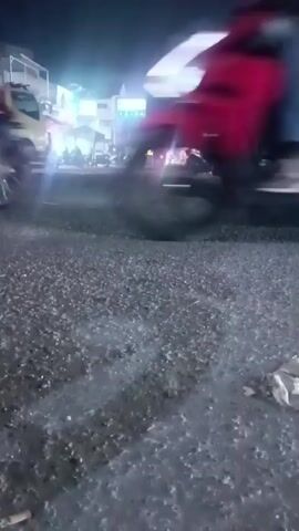 A low-angle, shaky video captured at night shows a busy intersection with several vehicles and people. In the foreground, a motorcycle with at least two occupants, one of whom is wearing a light-colored shirt and jeans, speeds past, its front wheel blurred due to motion. The road surface is dark and appears to be asphalt or gravel, with some tire marks visible. In the background, several other vehicles are present, including a large white truck with its headlights on, a smaller white vehicle, and other motorcycles. People are seen near the vehicles, and the scene is illuminated by artificial lighting from the vehicles and possibly streetlights. The overall impression is one of a chaotic and active nighttime scene.