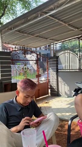 The video shows two individuals sitting outdoors under a covered patio. In the foreground, a person with short, dyed pink hair is looking down at a pink smartphone, holding it with both hands. They are wearing a dark polo shirt and light-colored pants. To their right, there is a clear plastic cup with a pink straw and a pink object, possibly another cup or container, also with a pink straw. In the midground and background, a second person is visible, wearing a white t-shirt and dark shorts, also looking at a smartphone they are holding with both hands. This person is holding a cigarette in their right hand. A white motorcycle is parked behind them, and a metal gate with decorative ironwork is visible in the background, leading to a building. The ground appears to be dirt and paved tile. The lighting suggests it is daytime, with shadows cast by the patio roof.