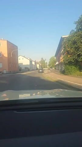 The video shows a view from inside a vehicle driving down a street. The street is lined with buildings on both sides. On the left are two-story buildings, one with an orange facade and another white building. On the right is a longer, multi-story building with a red facade and shops on the ground floor. A dark-colored car is driving ahead of the vehicle. The sky is clear and blue, indicating it is daytime. Trees are visible on the right side of the street, with some foliage extending over the sidewalk. There are streetlights and traffic signals along the road. The view is focused on the road ahead and the surrounding buildings.