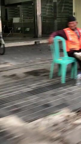 The video depicts a person sitting on a teal plastic chair. They are wearing a dark purple long-sleeved shirt, black pants, and white sandals. Over their shirt, they have an orange high-visibility vest. Their left hand is wrapped in a white bandage. The person is looking off to the left of the frame.

Beside the person, there is a bright orange traffic cone with a red base. The cone is positioned on a paved surface made of rectangular grey stones. In the background, a blurry figure and a building or structure are visible. The lighting suggests it could be daytime, possibly late afternoon, given the ambient light. The scene appears to be outdoors, possibly near a street or walkway. The overall impression is of a person taking a break in an urban or semi-urban environment.