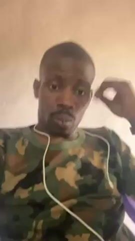 A man with dark skin, a short haircut, and a mustache is centered in the frame. He wears a camouflage t-shirt and white earbuds. He is sitting down and looks directly at the camera with a serious expression. He touches his nose and then rests his chin in his hand, looking thoughtful. He then points to his forehead. The background is a plain, light-colored wall.