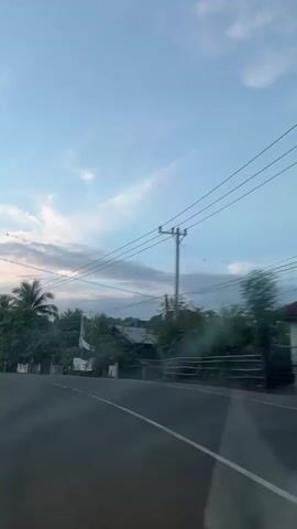 The video shows a view from a vehicle driving on a road. The sky is blue with some clouds, and the sun appears to be low on the horizon, suggesting either dawn or dusk. Palm trees and other vegetation line the side of the road, along with some buildings and utility poles. A white bus with illuminated lights is seen driving on the road, and a motorcycle is also visible. The road has white lane markings. The overall scene depicts a rural or suburban environment.