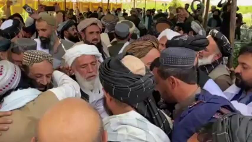Here is a concise description of the video:

The video shows a crowded scene with a large group of men. Many of them are wearing traditional clothing, including turbans and vests. Some individuals are hugging, suggesting a gathering of people who know each other or are celebrating an occasion. The background shows what appears to be an outdoor setting with a tent-like structure overhead. Several people are holding up phones, possibly recording or taking photos.