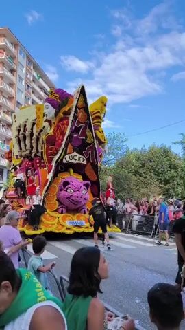 A colorful parade float, adorned with flowers and depicting a gambling theme, moves down a street. The float features a large, stylized pig with a mischievous grin, surrounded by what appear to be poker chips and cards. A queen figure is also prominent on the float, alongside other figures and imagery related to games of chance, including a "lucky" sign and a roulette wheel. People are watching the parade from the sidewalks, with some children visible in the foreground. The scene takes place on a sunny day with a blue sky dotted with clouds, and buildings line the street.